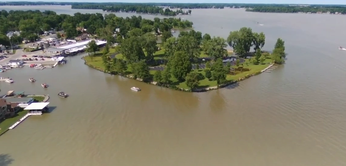 Panoramic daytime view of sparkling Indian Lake from Russells Point, Ohio, featuring the Sandy Beach Bridge connecting to a green shoreline, with recreational boats on the water and the village in the distance.